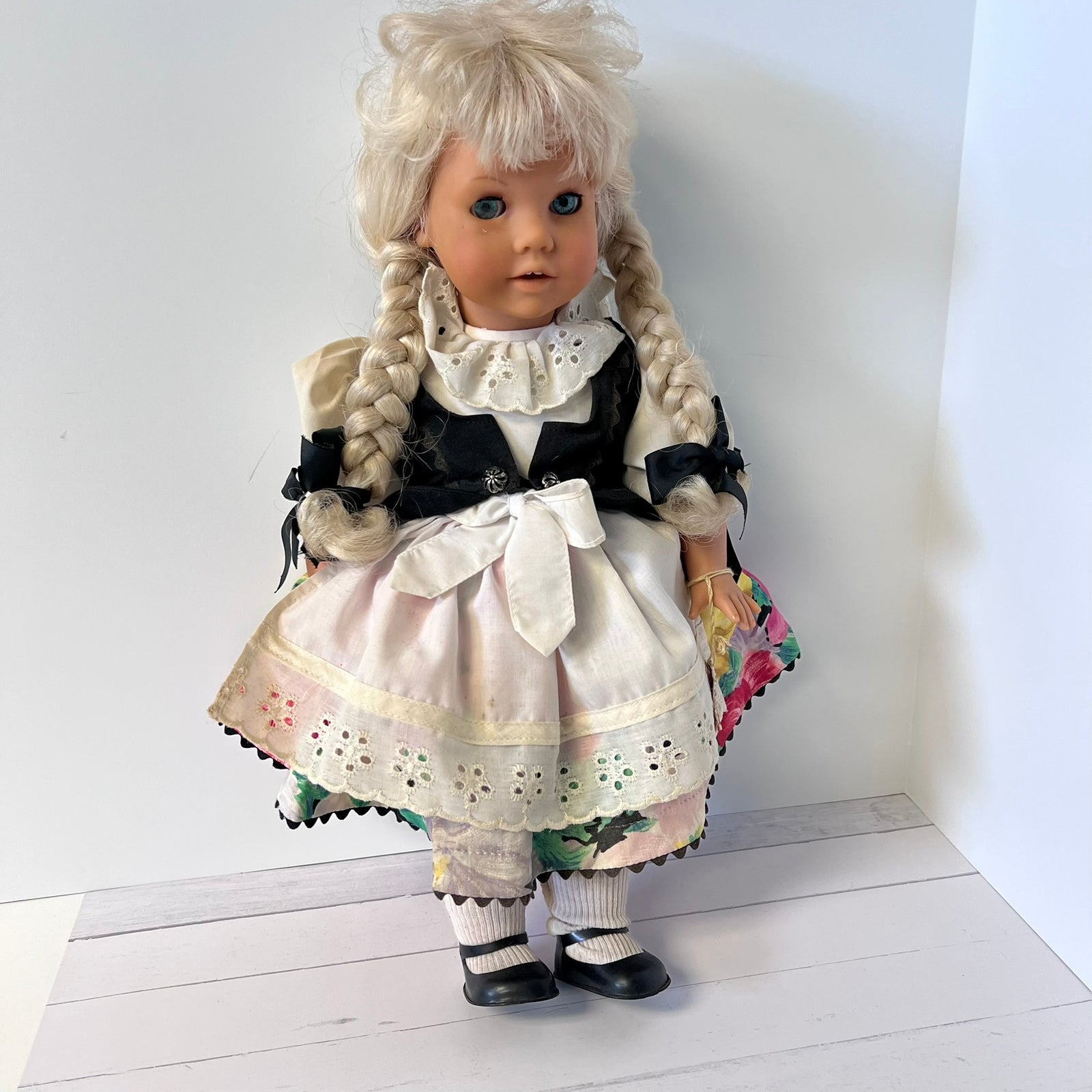 A vintage-style porcelain doll with long blonde pigtails, wearing a black and white dress with a large white bow, standing against a plain white wall.