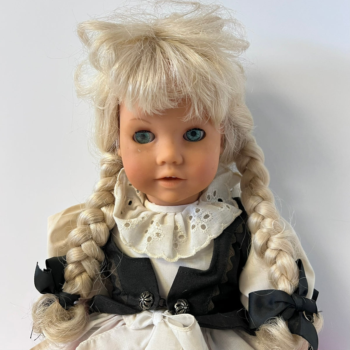A vintage-style blonde doll with blue eyes, styled with a messy wig, braided pigtails, and a black and white dress with a lace ruffle collar, photographed against a plain light gray background.