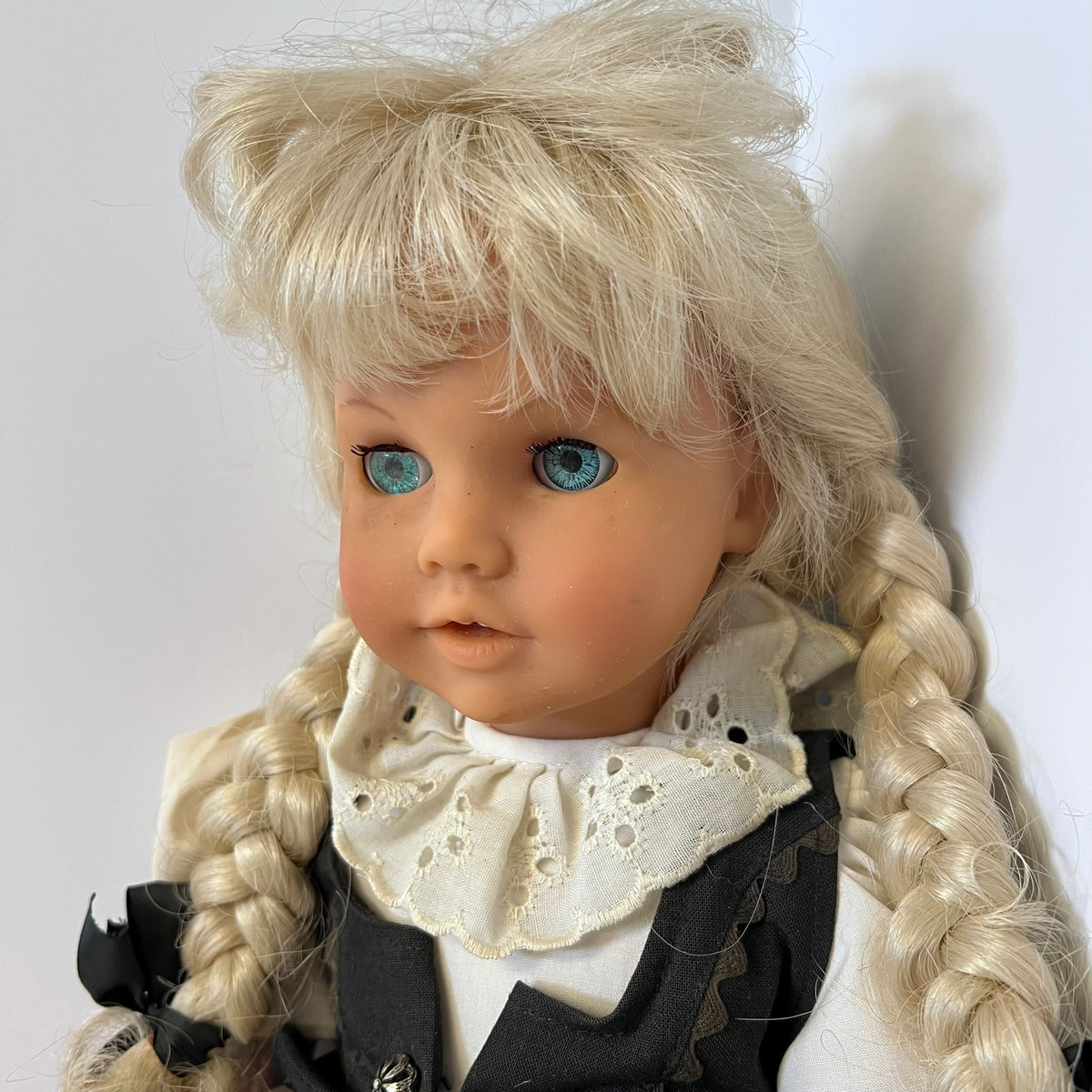 A close-up of a vintage-style blonde doll with blue eyes, wearing a white lace ruffled collar, a white top, and a dark gray pinafore dress. The doll has long braided hair and is photographed against a plain light background.