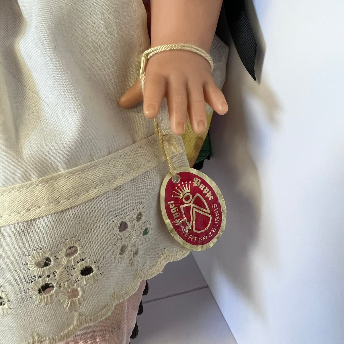 A close-up of a doll's hand wearing a beige braided bracelet, holding a round red and gold hanging tag that reads 'Happy Birthday' and 'Pia's First Birthday'.