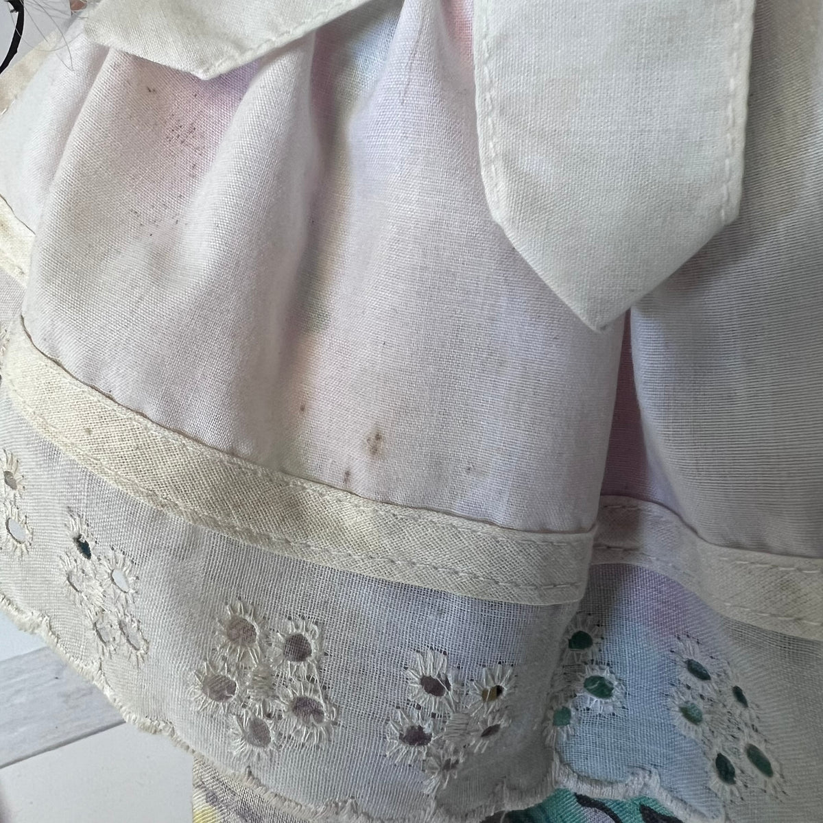 A close-up of a vintage white or off-white cotton dress or skirt fabric, featuring a pointed collar, a decorative band with floral eyelet embroidery, and a soft pastel rainbow sheen over the material.