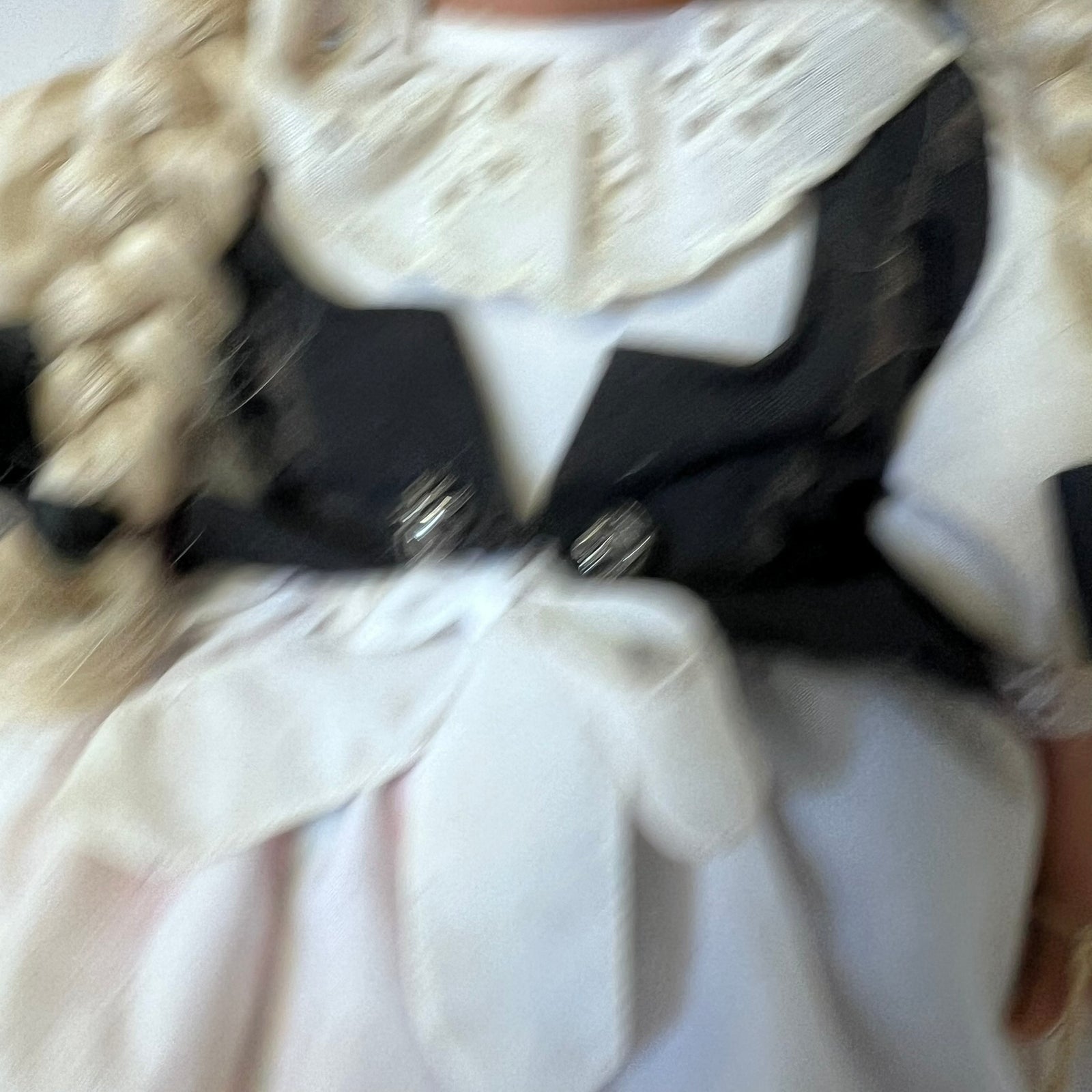 A close-up of a doll or toy figure wearing a black and white outfit with a large white lace ruffled collar and a white bow at the waist.