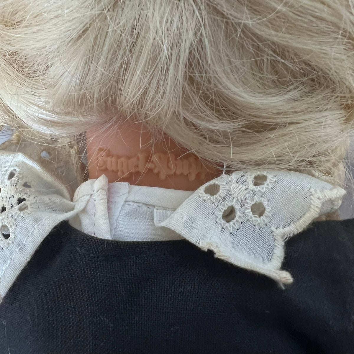 Close-up back view of a doll with platinum blonde hair, a beige neck, a white lace collar, and a black dress.