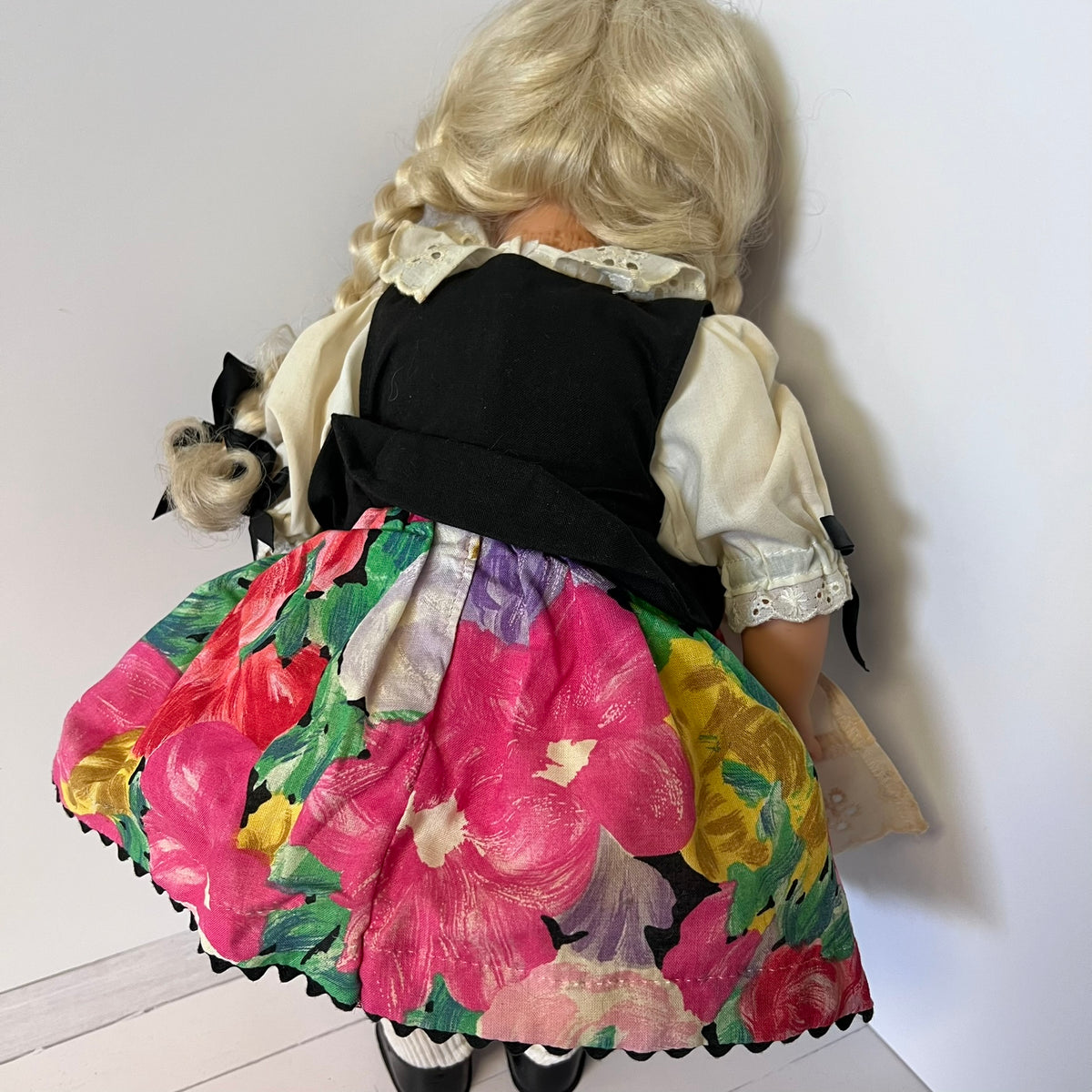 A vintage-style doll viewed from behind, wearing a black vest, cream lace-trimmed blouse, and a vibrant floral skirt with pink, yellow, green, and purple flowers.