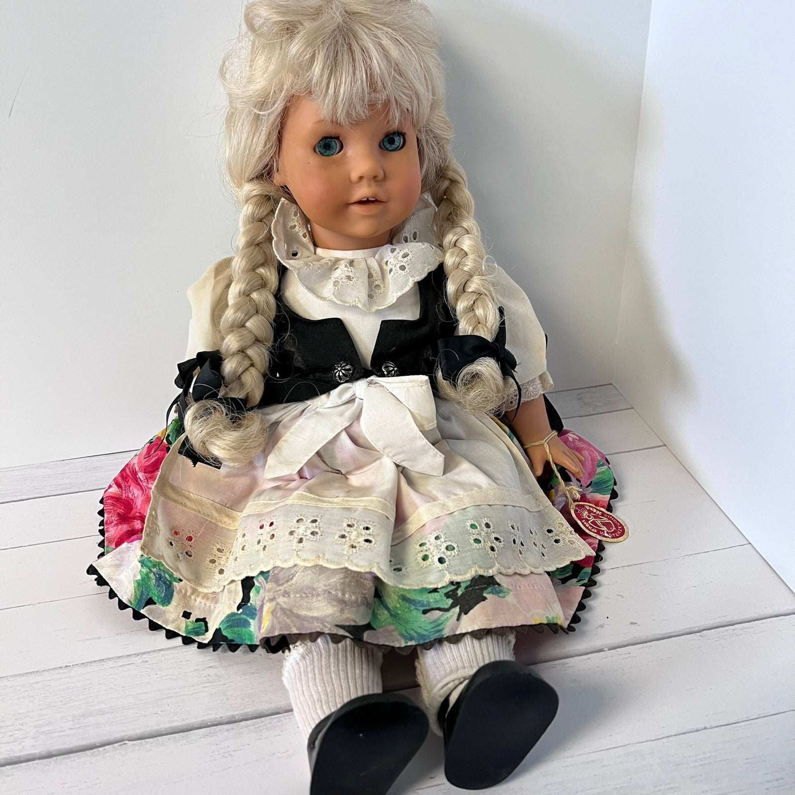 A vintage-style doll with long blonde pigtails sits on a white wooden floor against a white wall. The doll is wearing a black and white dress with a floral multicolor skirt, white ribbed socks, and black shoes.