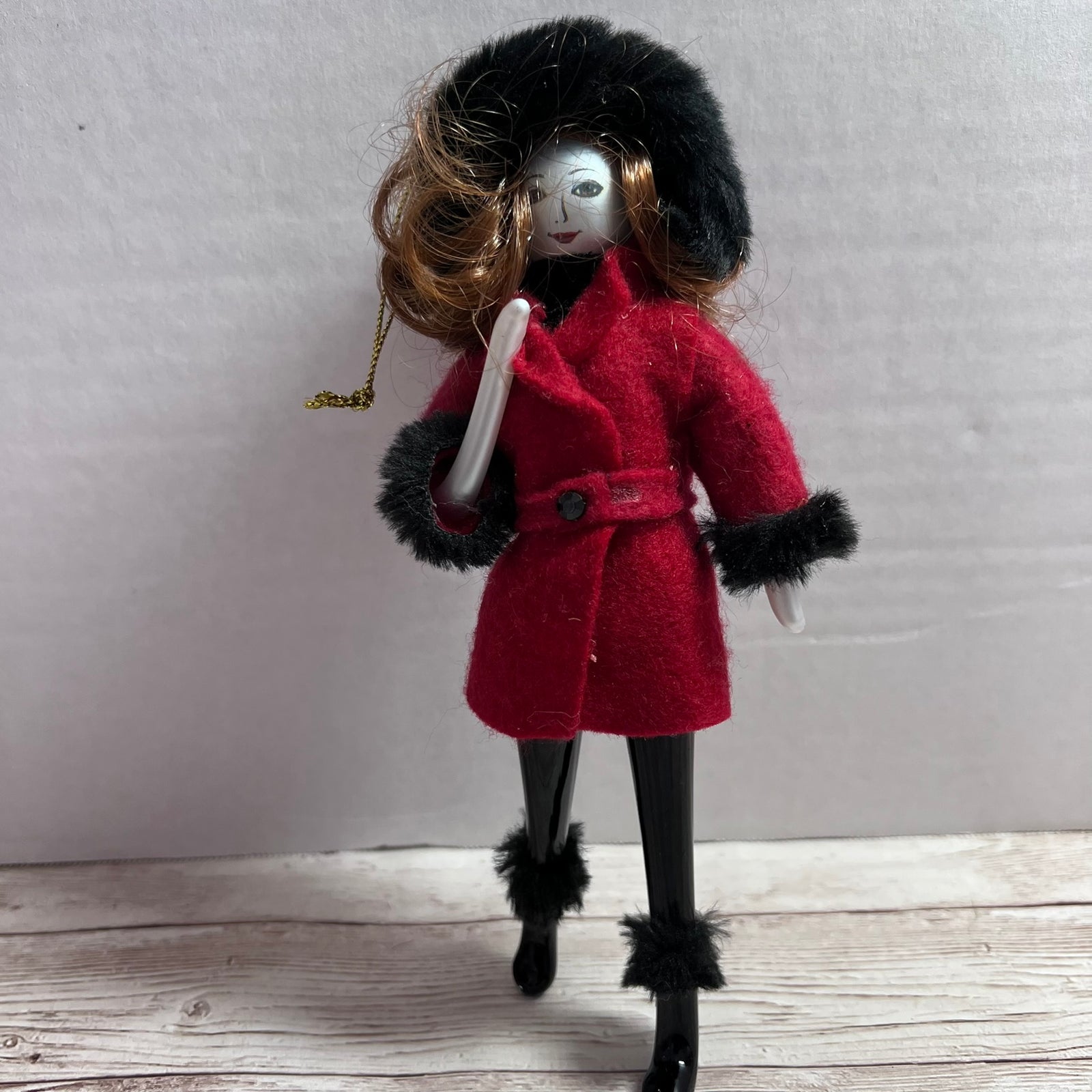 A vintage-style doll with long brown hair wearing a bright red coat with black fur trim, black tights, and a black fur hat, standing on a light wood surface against a plain light gray wall.
