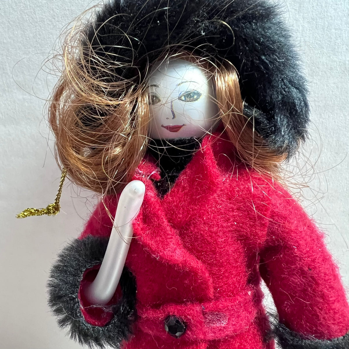 A vintage-style doll with a white face, brown hair, a black fur hat, and a bright red coat, holding a white object against a plain light background.