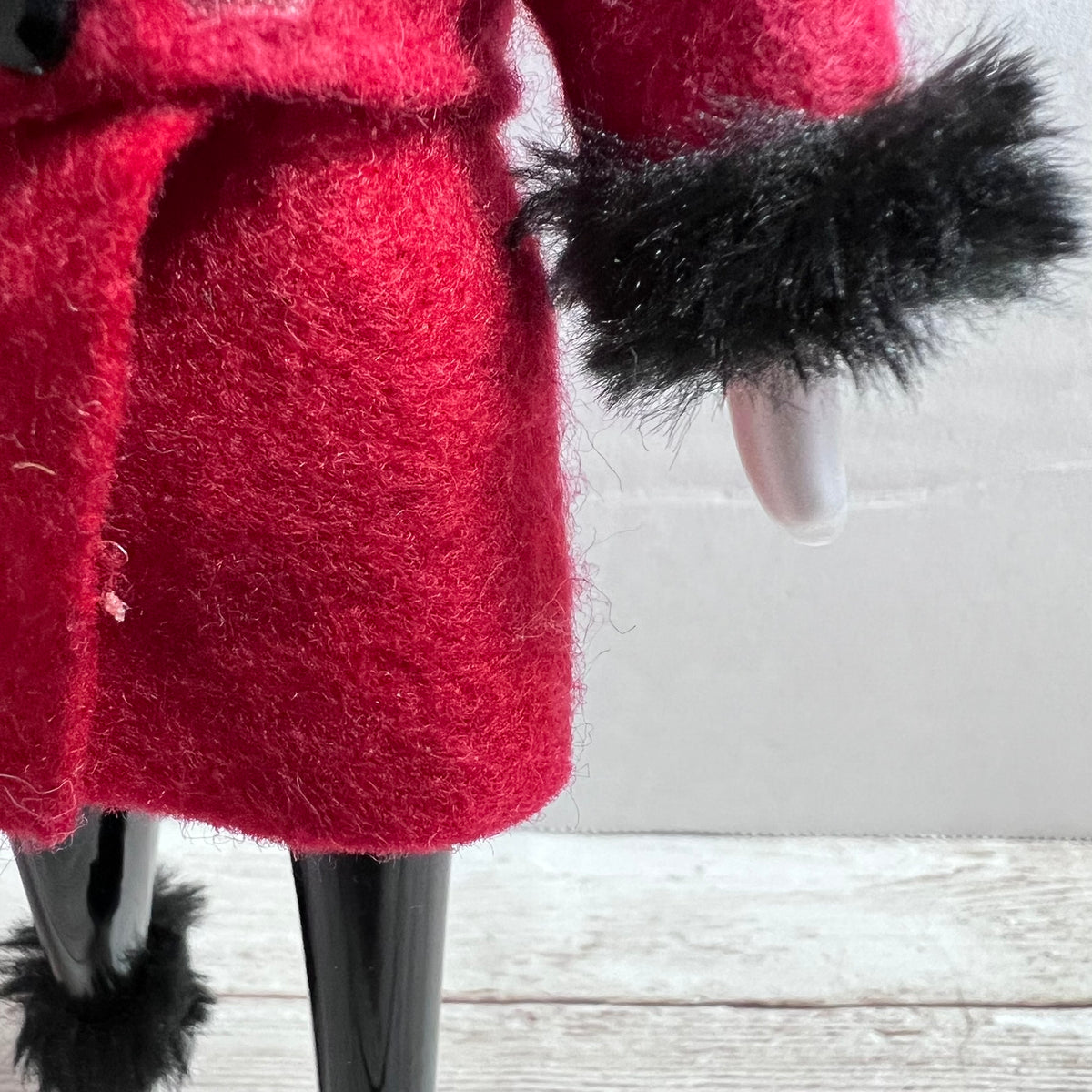 A close-up of a red fuzzy coat with black faux-fur trim and black legs, shown against a light gray wall and white wood floor.