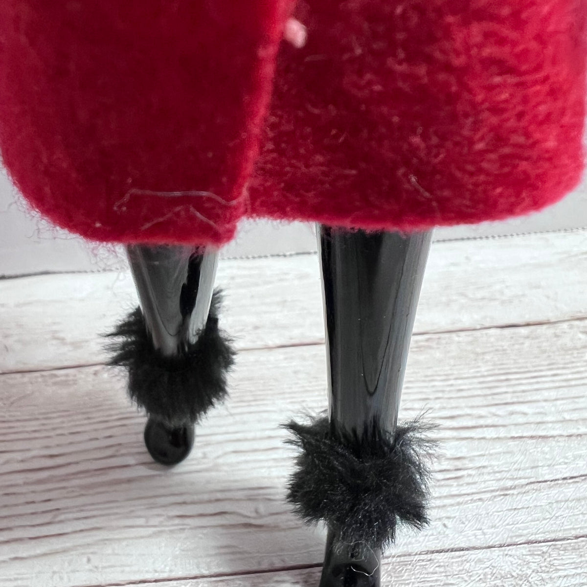 A close-up of a red fuzzy garment, likely a skirt or coat hem, hanging over glossy black legs with black faux-fur trim near the bottom, set against a light wood floor background.