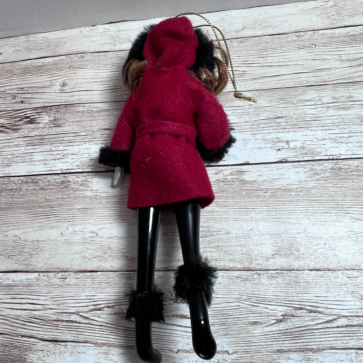 A doll viewed from the back wearing a bright red hooded coat with black faux-fur trim, black pants, and black boot covers, standing against a whitewashed wood plank background.