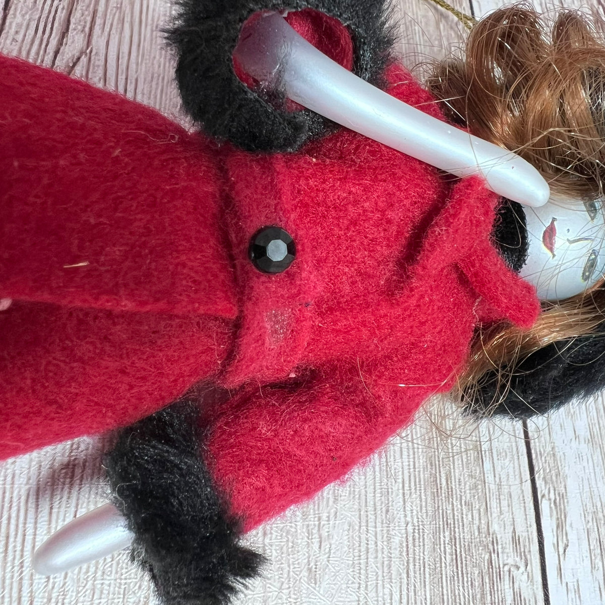 A close-up of a handmade doll or plush figure wearing a bright red felt coat with black trim and a black button, lying on a light wood surface.