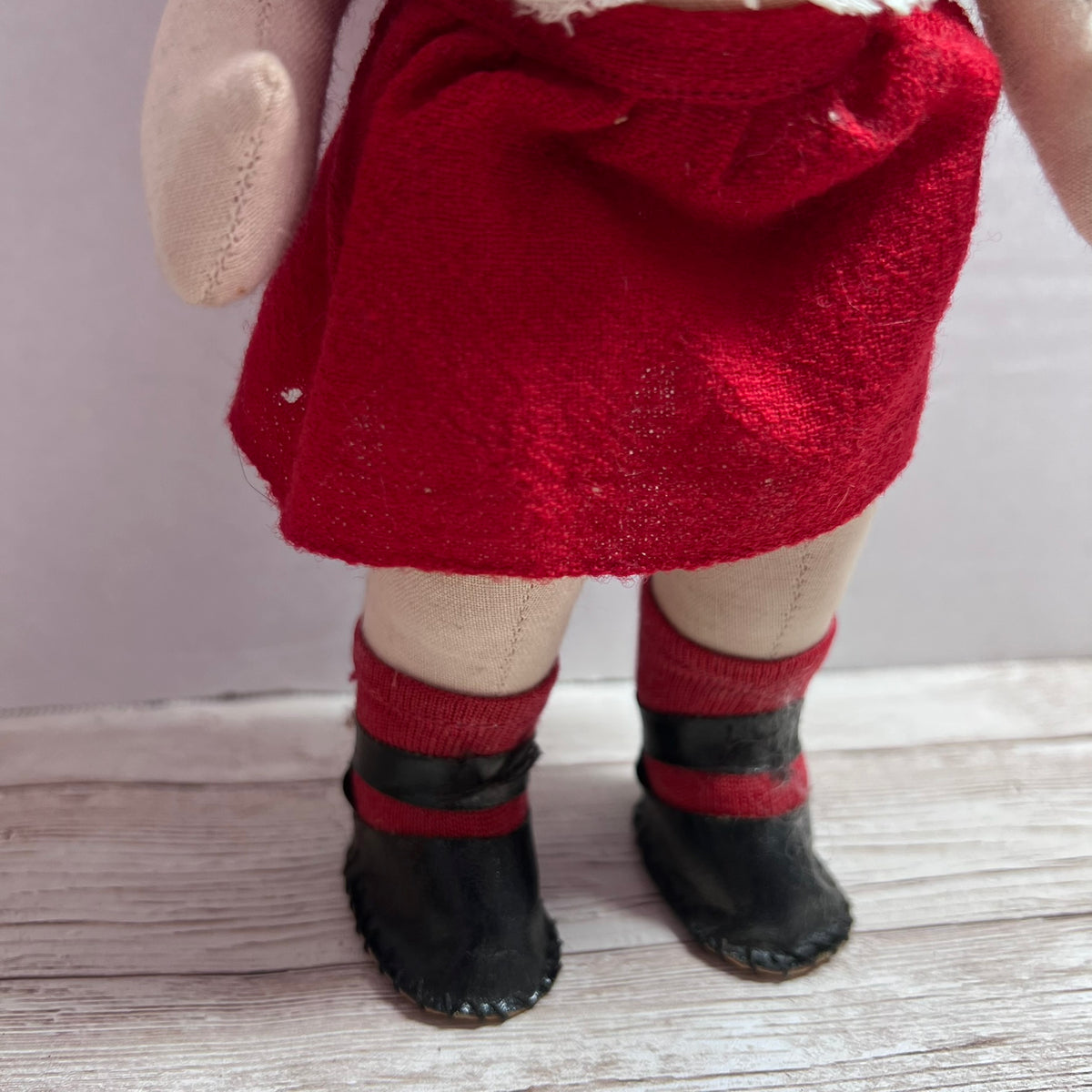 Close-up of a doll's lower body wearing a red skirt, beige fabric legs, red and black boot covers, and black booties, standing on a light wood surface.