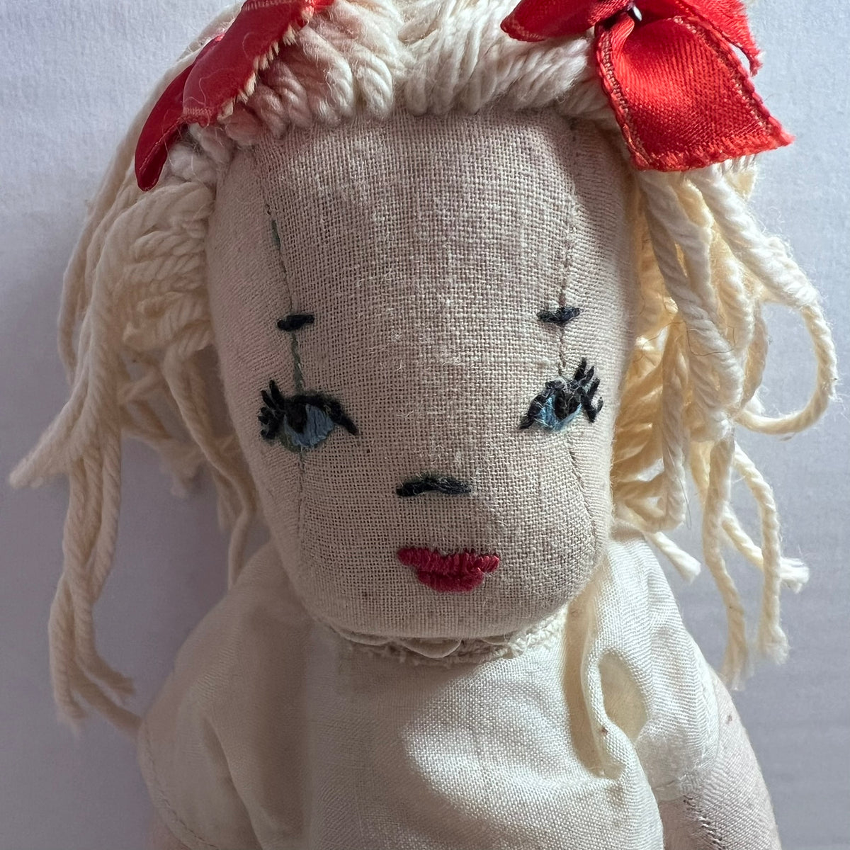 A vintage-style handmade fabric doll with a linen face, cream-colored yarn hair, and red ribbon bows, shown in a close-up shot against a plain light background.
