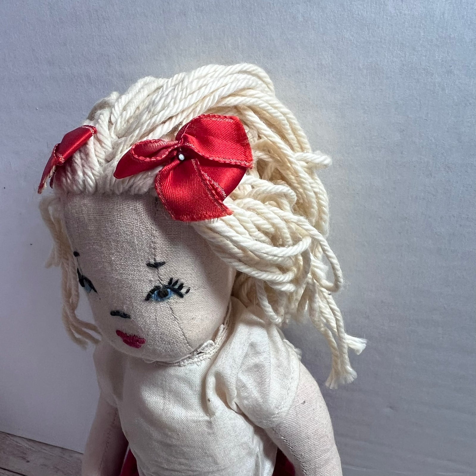 A handmade fabric doll with a cream-colored face and body, styled with curly off-white yarn hair adorned with two red satin bows, photographed against a plain light gray wall.