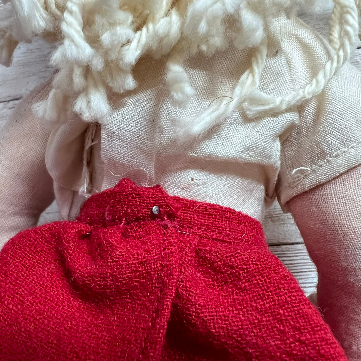 Close-up of a handmade fabric doll or rag doll showing cream-colored linen clothing, a bright red skirt, and textured off-white yarn hair, pinned together on a light wood background.