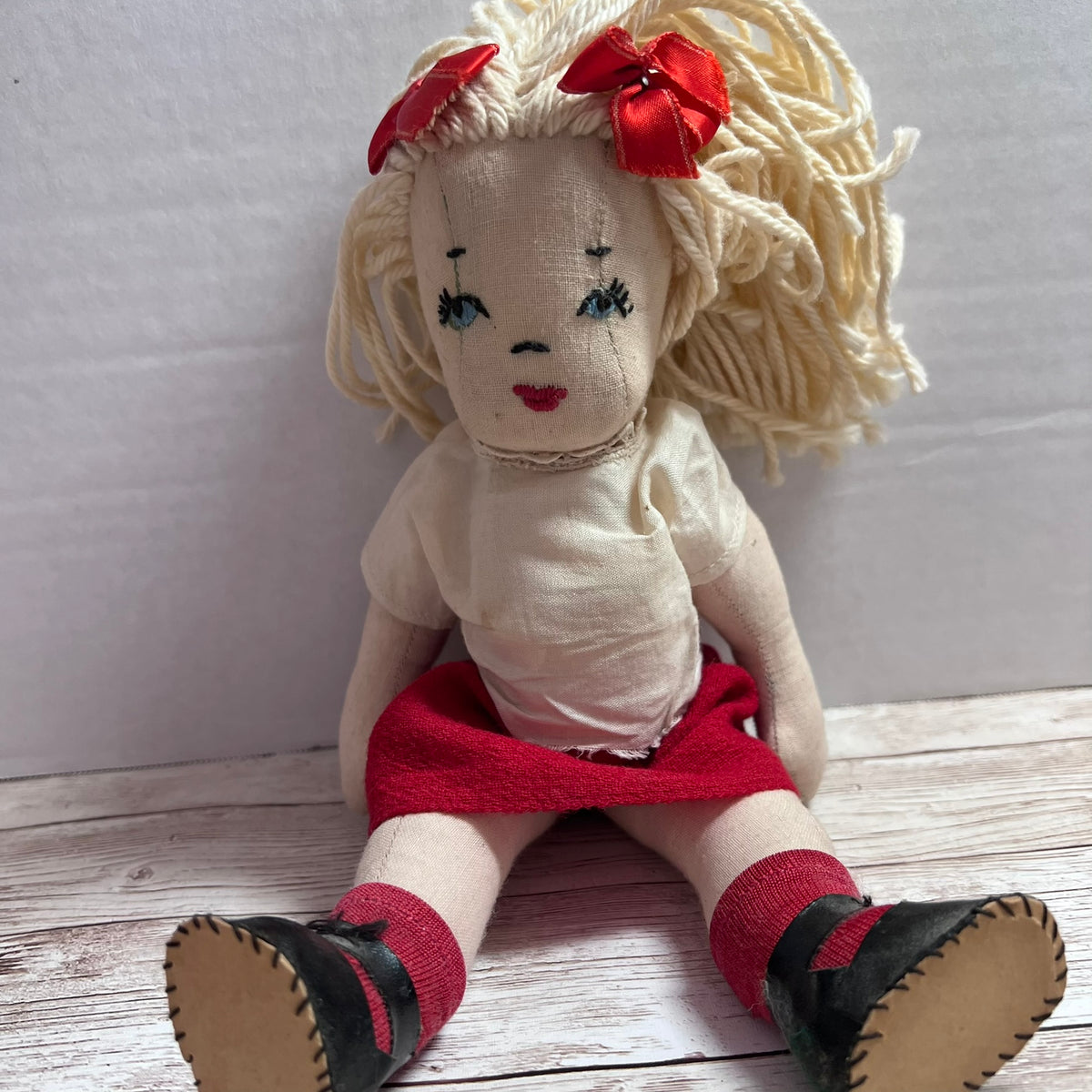 A handmade rag doll with cream-colored yarn hair, two red bows, a cream shirt, a red skirt, and black shoes, sitting on a light wood floor against a white wall.