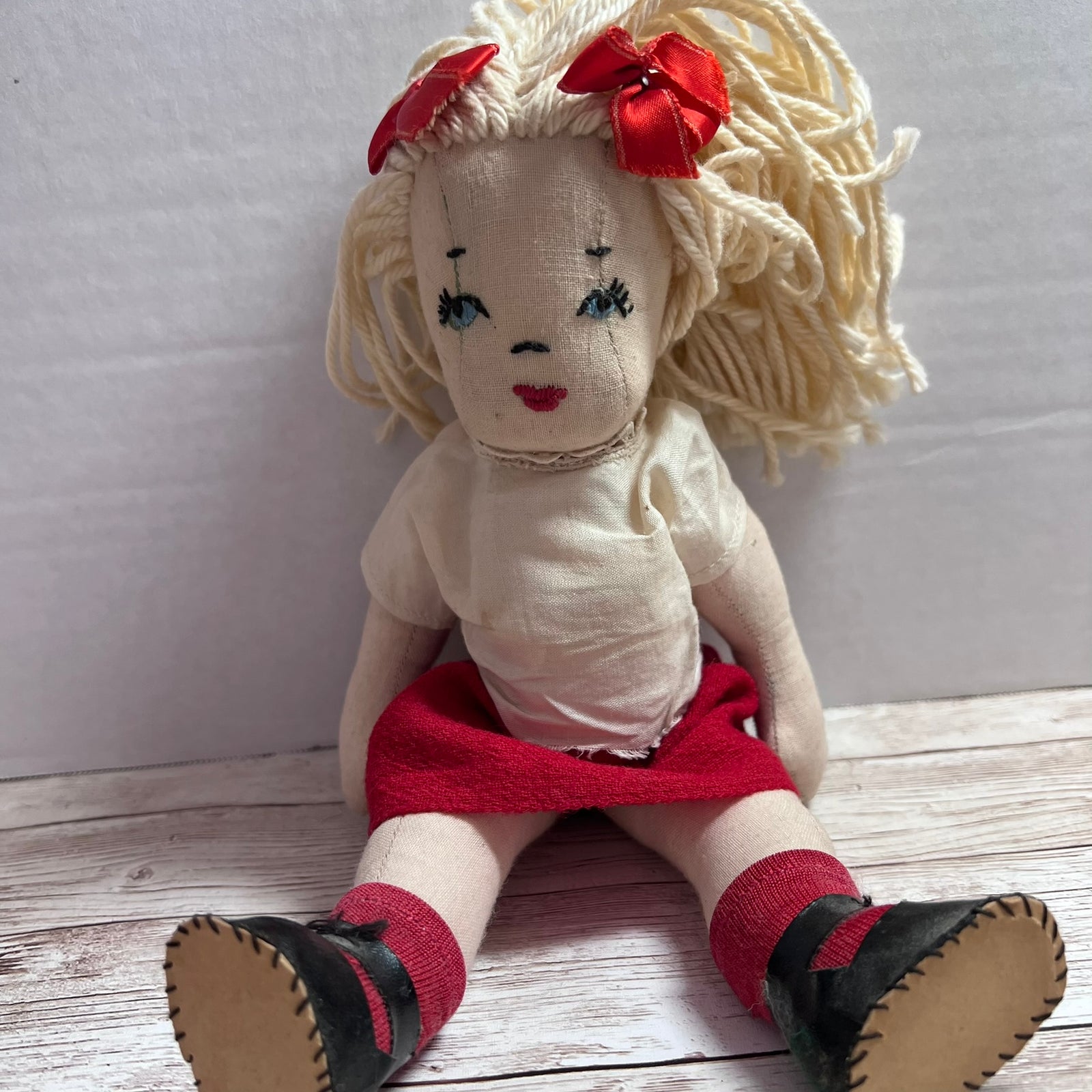 A handmade rag doll with cream-colored yarn hair, two red bows, a cream shirt, a red skirt, and black shoes, sitting on a light wood floor against a white wall.