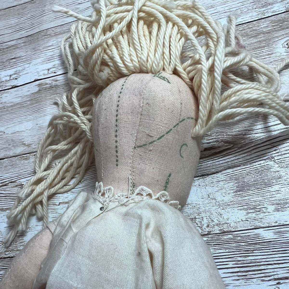 A handmade fabric doll with messy cream-colored yarn hair, stitched on a linen bodice, lying on a rustic white-washed wooden surface.