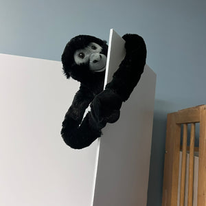 Black monkey plush toy peeking over a white surface with a gray wall in the background