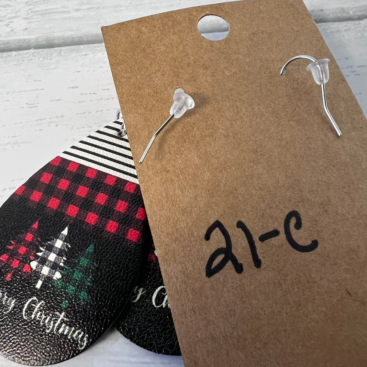 Pair of silver earrings on a brown card with '21-C' printed on it, next to Christmas-themed coasters.