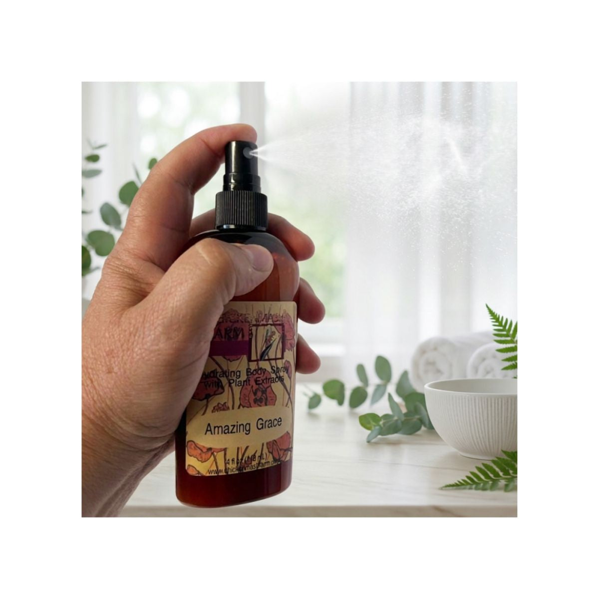 A hand holding a brown spray bottle labeled 'Amazing Grace' and spraying a mist, set against a softly blurred bathroom background with a white bowl, green leaves, and a bright window.