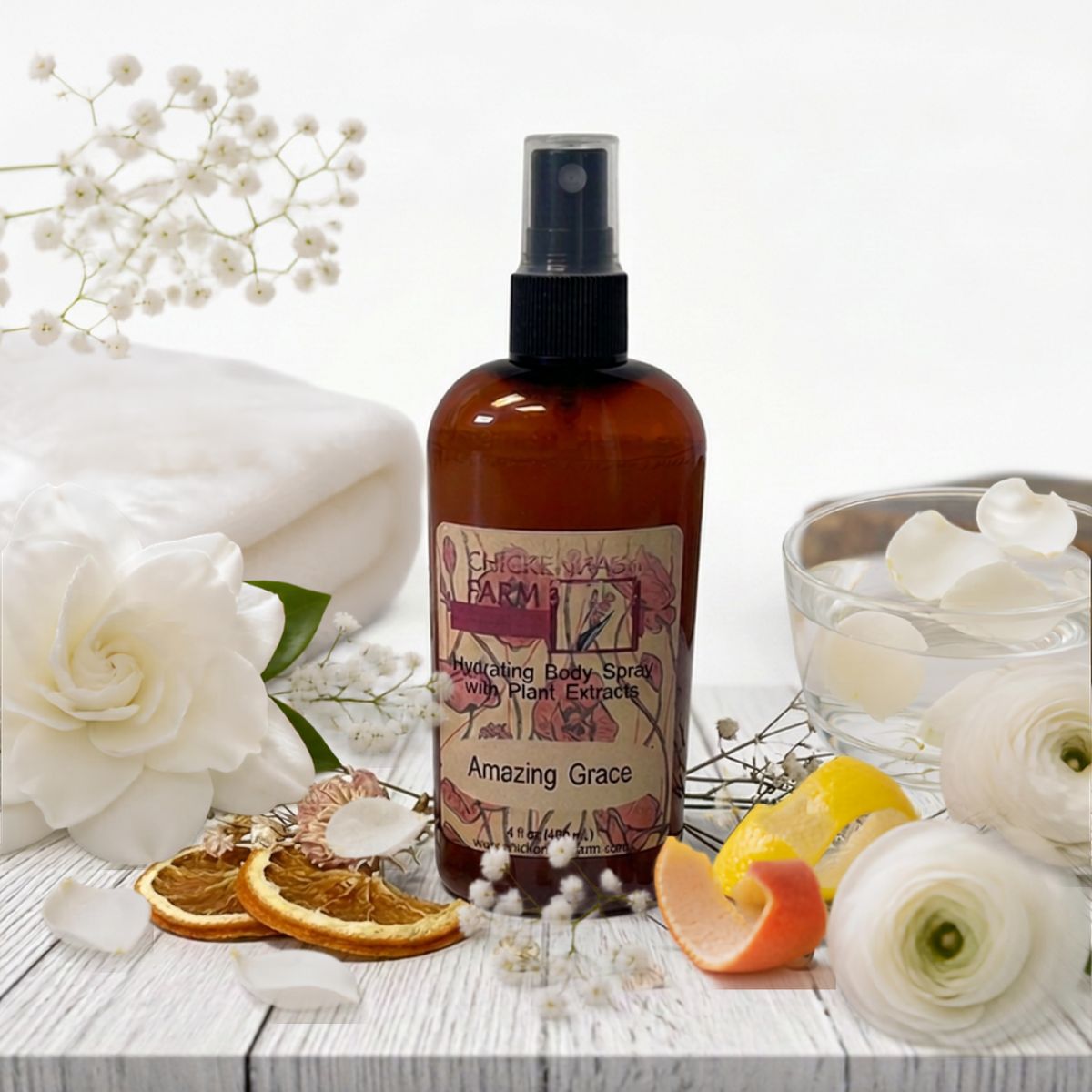 An amber spray bottle labeled 'Amazing Grace' with floral artwork on the front, styled on a white wooden surface with white flowers, dried orange slices, a folded white towel, and a glass bowl of petals.
