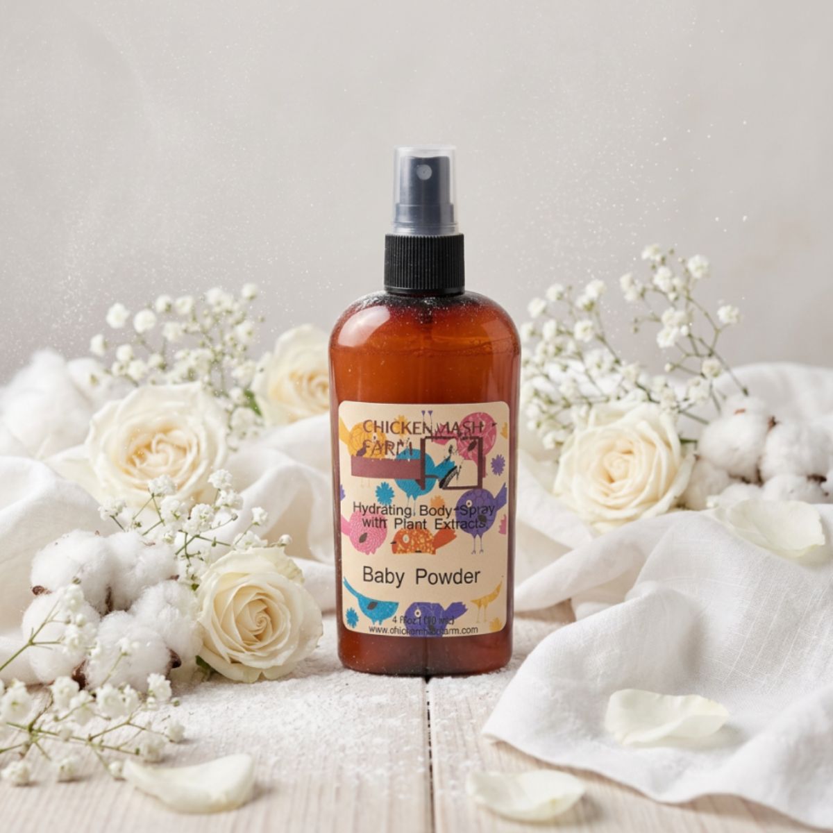 An amber 
spray bottle labeled 'Chickenmash Farms Baby Powder Hydrating Body Butter with Plant Extract' is styled on a light wood surface with white roses, cotton balls, baby's breath, and white fabric.