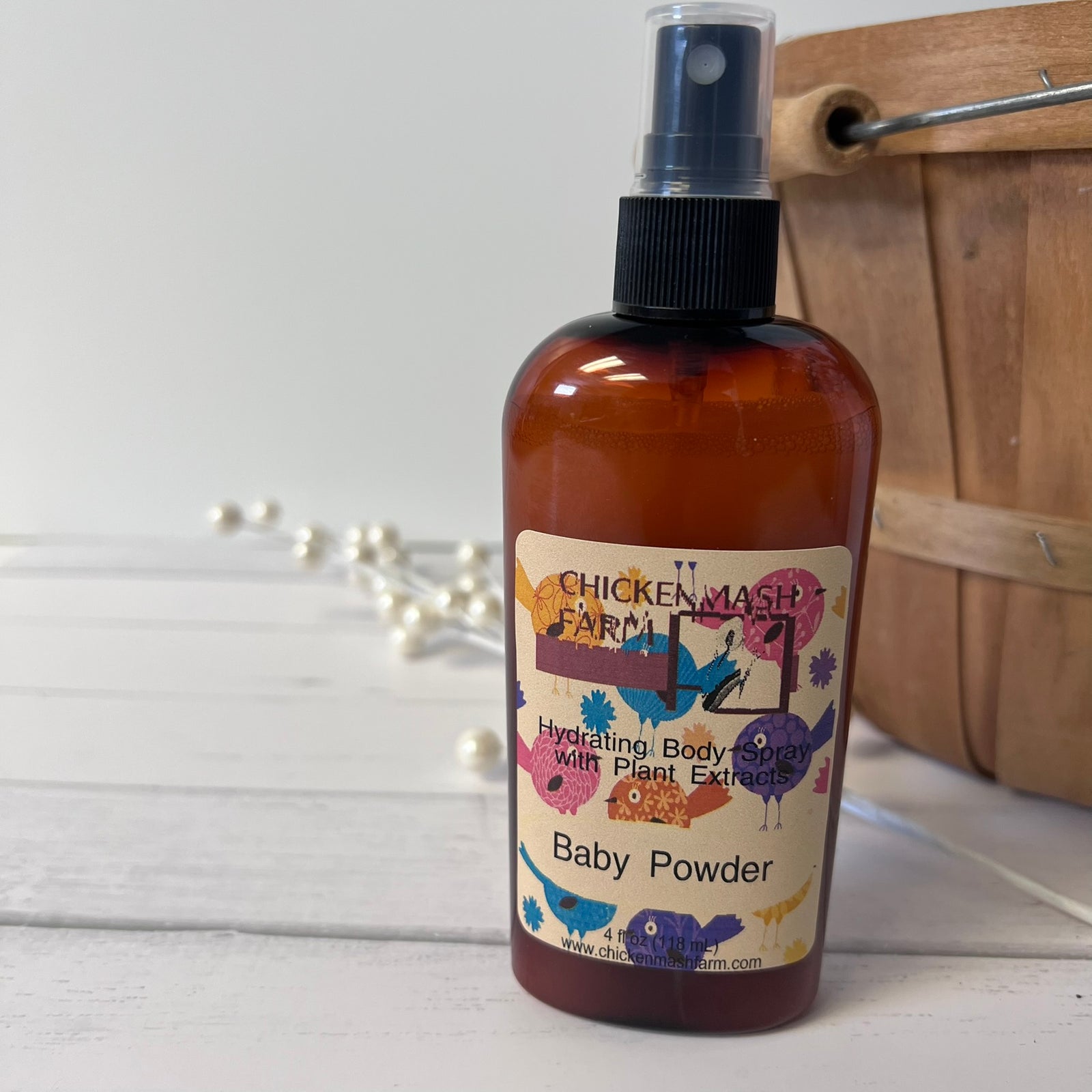 A bottle of Chicken Mash Farm Baby Powder Hydrating Body Soap with Plant Extracts, in an amber brown bottle with a black spray cap, set on a white wooden surface with a wooden basket and white pearls in the background.