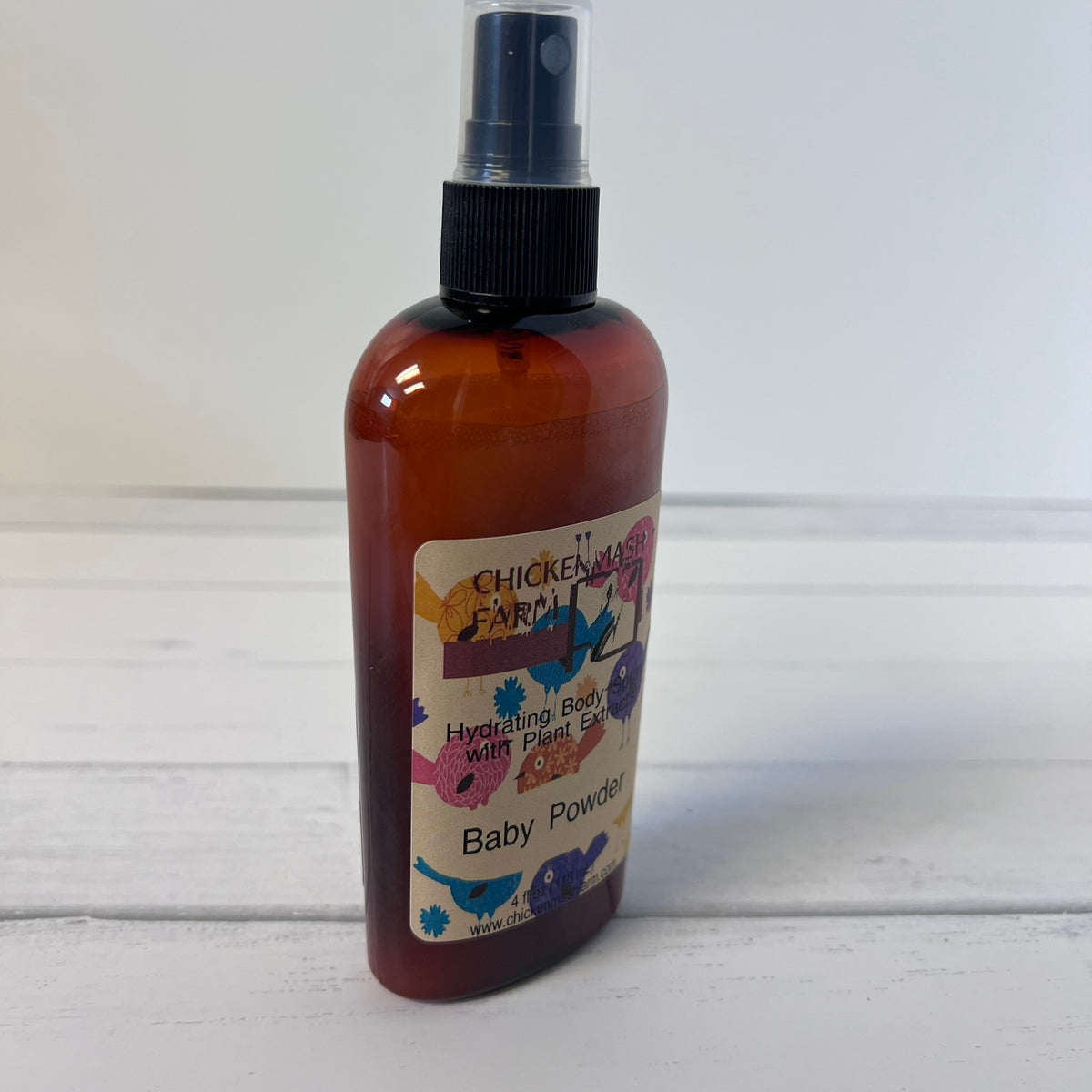 An amber plastic spray bottle with a black spray top, featuring a cream label with colorful cartoon animals and the text 'Chicken Farm Hydrating Baby Milk Plant Based Baby Powder'.