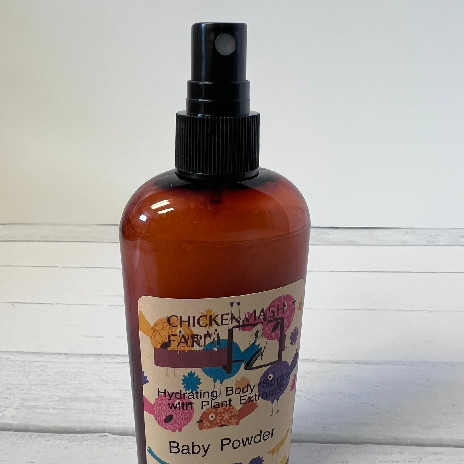 An amber plastic spray bottle of Chicken Mash Farm Baby Powder Hydrating Body wash with Plant Extract, featuring a black spray nozzle and a colorful label, set against a white wooden background.