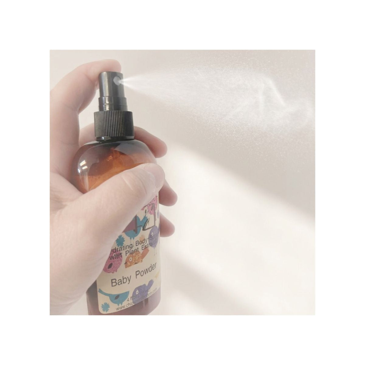 A hand holding a brown spray bottle labeled 'Baby Powder' and spraying a fine mist against a light beige background.