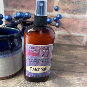 Bottle of Chicken Mash Farm Patchouli hydrating body spray on a wooden surface with blueberries in the background.