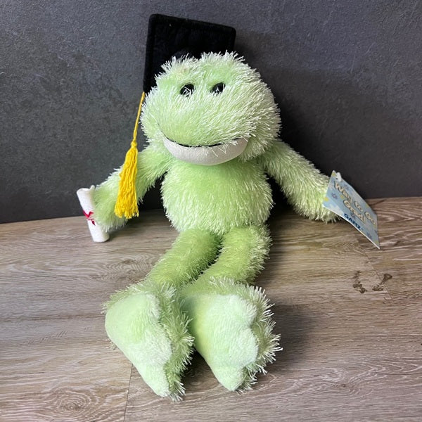 Green plush frog with a graduation cap and tassel on a wooden surface.