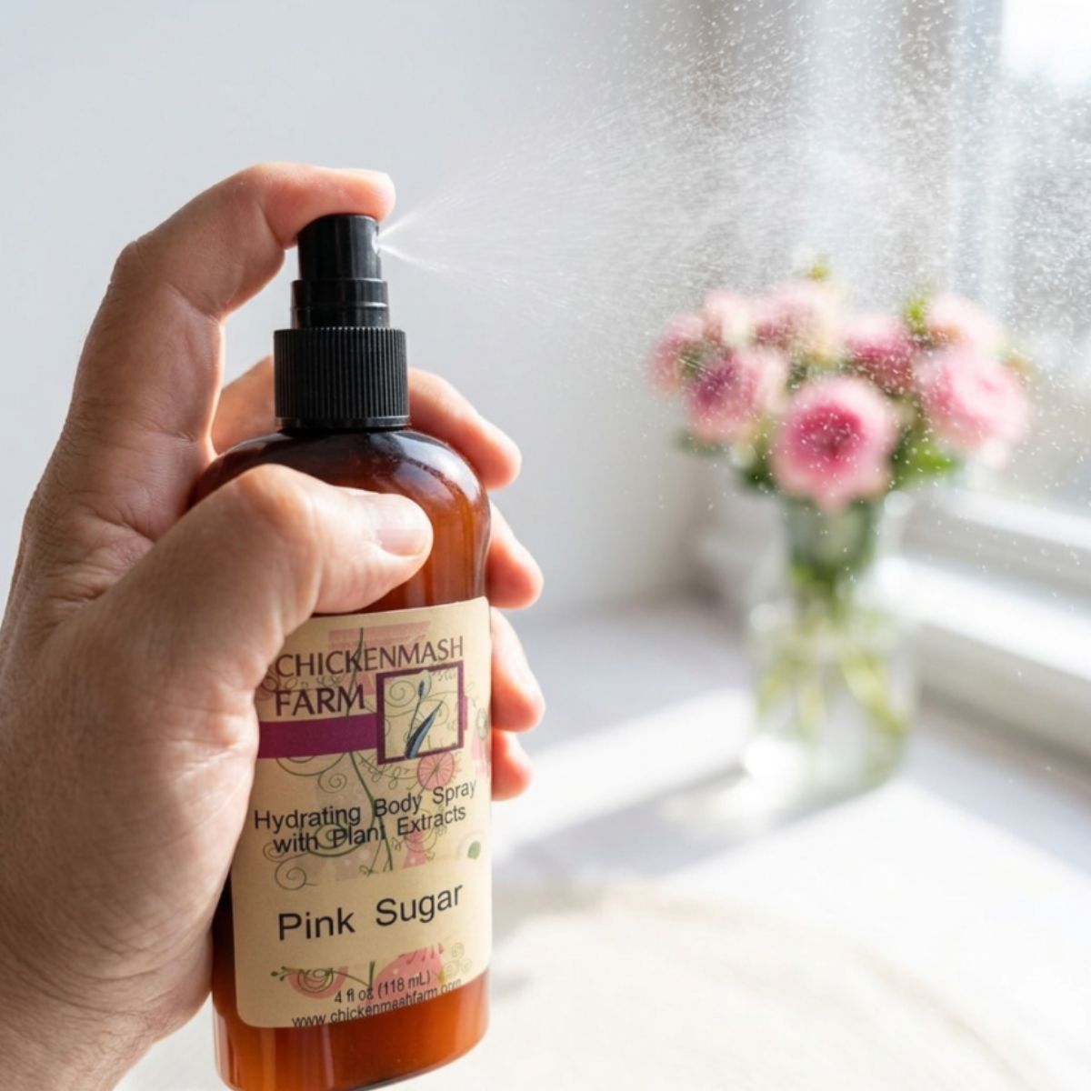 A hand holds a bottle of Chicken Mash Farm Hydrating Body Spray with Plant Extracts, Pink Sugar scent, spraying a fine mist in front of a softly blurred window with pink flowers.