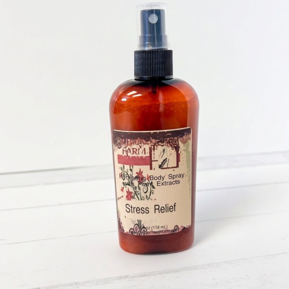 An amber body spray bottle with a black spray nozzle and a beige label reading 'Stress Relief' and 'Harvesting Body Spray Plant Extracts' by Chickenmash Farm, set against a white wood plank background.